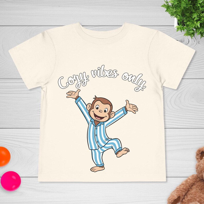 Op de afbeelding: Cr&egrave;mekleurig T-shirt met een cartoon aap in blauw-witte gestreepte pyjama met uitgestrekte armen. De tekst "Cozy vibes only" staat in een speels lettertype boven de aap. Een leuk en comfortabel T-shirt voor kinderen.