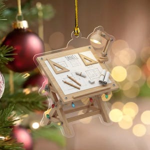 May include: A clear acrylic Christmas ornament shaped like a drafting table with a wooden frame. The table surface displays architectural blueprints with the name Daniel, two triangles, two pencils, a rolled paper, and an eraser. A small lamp is attached.