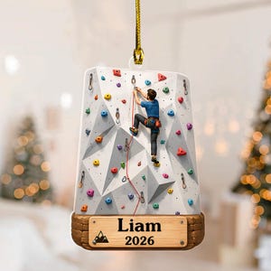 May include: A flat, rectangular hanging ornament featuring a white climbing wall with colorful holds and a climber in a blue shirt and dark pants. A wooden base at the bottom displays the name Liam and the year 2026 with a mountain icon.