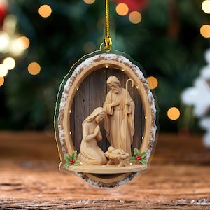May include: A Christmas ornament featuring a Nativity scene. The ornament has a wooden frame with a bark-like edge and a gold hanging string. Inside, a carved wooden depiction of Mary, Joseph, and baby Jesus is visible, with holly accents.