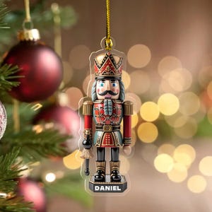 May include: A flat, acrylic nutcracker ornament featuring a soldier in a red, blue, and gold uniform with a black mustache. The figure wears a tall crown and stands on a black base with the name DANIEL printed in white capital letters.