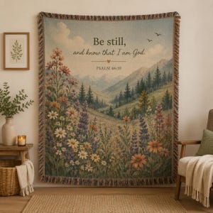 May include: A woven throw blanket featuring a landscape illustration of a mountain valley with wildflowers in the foreground. The text reads, Be still, and know that I am God. PSALM 46:10. The blanket has a fringed edge and a muted color palette.