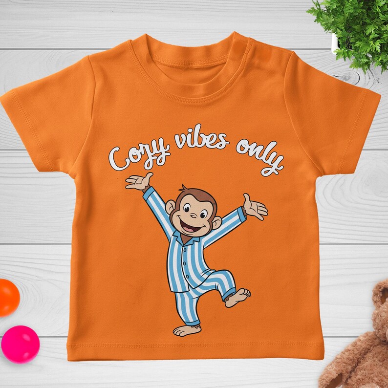 Op de afbeelding: Oranje peuter-T-shirt met een cartoon aap in blauw-witte gestreepte pyjama met uitgestrekte armen. De tekst "Cozy vibes only" staat in sierlijke letters boven de aap. Het shirt is gemaakt van zacht materiaal.