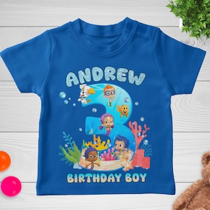 May include: A blue t-shirt featuring the name "ANDREW" in glittery, light blue letters above a large number "3" with cartoon characters. The words "BIRTHDAY BOY" are printed below the number. The shirt has a crew neck.