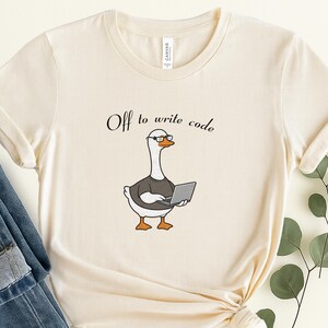 May include: A cream-colored crew neck t-shirt featuring a graphic of a white goose wearing black glasses and a dark gray shirt while holding a silver laptop. Above the goose, the text reads, Off to write code, in a black cursive font.