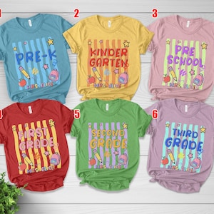 May include: Six colorful t-shirts with school-themed designs. Each shirt features a grade level: Pre-K, Kindergarten, Preschool, First Grade, Second Grade, and Third Grade. Designs include pencils, apples, stars, and backpacks.