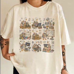 May include: Cream-colored t-shirt featuring a whimsical graphic of a duck in a plaid shirt, engaged in various STEM activities. The design includes lab equipment, a rocket, and a laptop, creating a playful and educational theme.