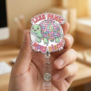 May include: A badge reel with a cartoon turtle design. The turtle has a green body and a disco ball shell. The text on the badge reel says "Less Panic, More Disco" in a playful font. The reel has a clear plastic clip and a retractable cord.