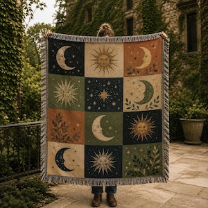 May include: A woven throw blanket featuring a grid of twelve rectangular panels displaying celestial motifs including suns, crescent moons, stars, and leafy branches in a palette of navy blue, sage green, cream, and terracotta with fringed edges.