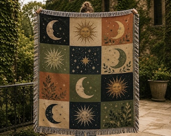 May include: A woven throw blanket featuring a grid of twelve rectangular panels. Each panel displays celestial motifs including suns with faces, crescent moons, stars, and leafy branches in a palette of navy blue, olive green, and terracotta.