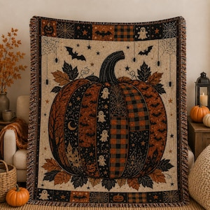 May include: A woven throw blanket featuring a large central pumpkin design in orange and black. The pumpkin is decorated with ghosts, bats, stars, and plaid patterns. The border includes spiderwebs, jack-o'-lanterns, and autumn leaves.