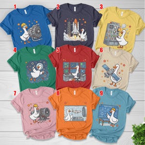 May include: A collection of nine colorful t-shirts, each featuring a cartoon goose in various space-themed scenarios. Designs include the goose operating machinery, near a rocket launch, and working at a control panel. The shirts are in various colors.