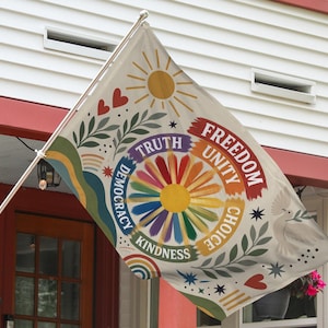 May include: A decorative flag with a cream background, featuring a colorful floral design with the words "Freedom, Unity, Choice, Kindness, Truth, Democracy". The flag also includes a sun, hearts, and a dove with an olive branch.