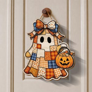 May include: A ghost-shaped wall hanging featuring a patchwork design of orange, blue, and cream floral and plaid patterns. The ghost has a large bow at the top and carries an orange jack-o'-lantern bucket filled with candy corn and leaves.