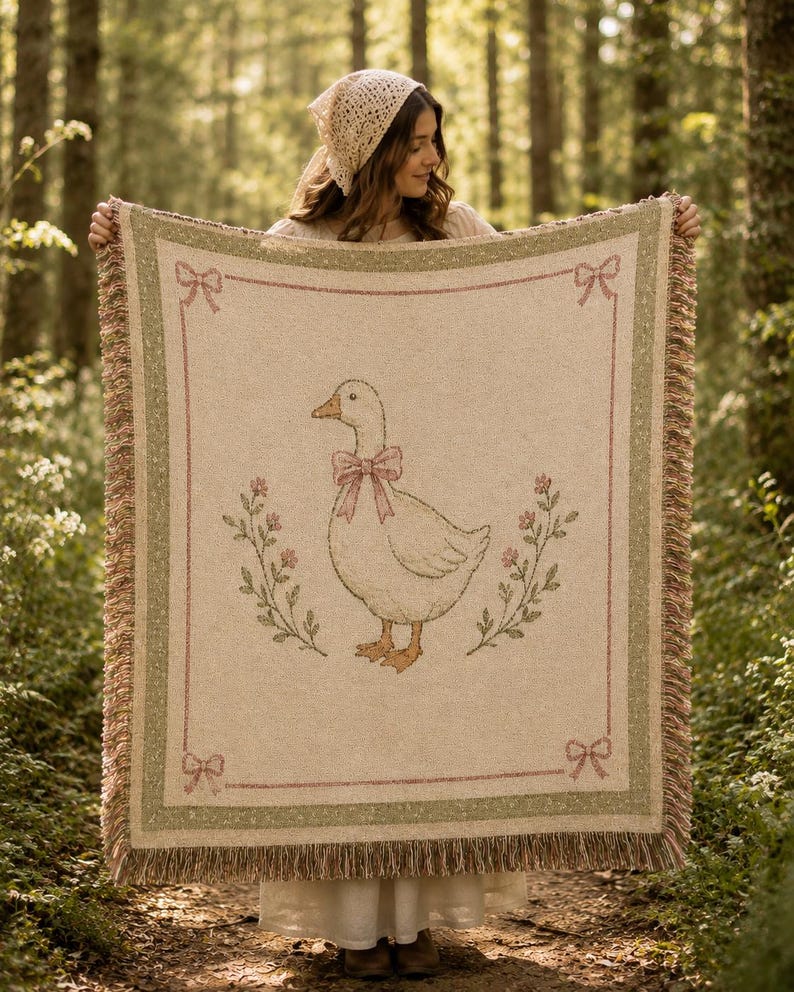May include: A rectangular woven throw blanket featuring a central illustration of a white goose wearing a pink bow. The design includes floral sprigs on either side, a pink border with corner bows, and a green outer frame with fringed edges.