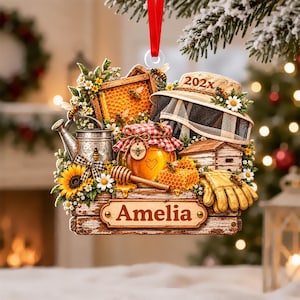 May include: A decorative hanging ornament featuring a wooden crate base with the name Amelia printed on it. The design includes a honey jar, a honeycomb, a beekeeper hat with 202x text, a metal watering can, sunflowers, and bees.