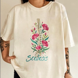 May include: Cream-colored t-shirt featuring a spine illustration adorned with pink flowers and green leaves. The word "Scoliosis" is written in teal script below the floral design. The shirt has short sleeves.