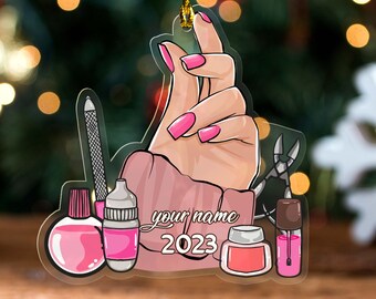 May include: A clear acrylic Christmas ornament featuring a hand with pink nails, nail polish bottles, and nail tools. The text "your name 2023" is also included. The ornament is suspended by a gold string.