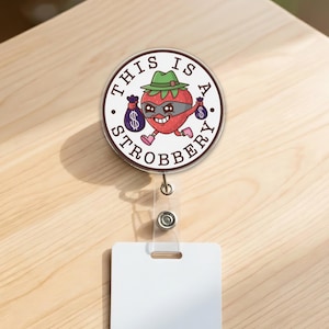 May include: A round badge reel with a cartoon strawberry wearing a mask and hat, holding money bags. The text "THIS IS A STROBBERY" is printed around the image. The reel has a clear plastic strap and a white card holder.