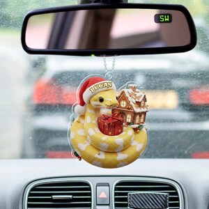 May include: A yellow and white snake-shaped Christmas ornament with a Santa hat that says "LUCAS". The ornament is holding a red gift box and is next to a gingerbread house. The ornament is hanging from a car's rearview mirror.