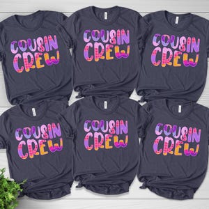 May include: Six dark charcoal gray short-sleeve t-shirts arranged in two rows. Each shirt features the text "COUSIN CREW" printed in a bold, multi-colored font with purple, pink, and orange patterns including polka dots and plaid designs.