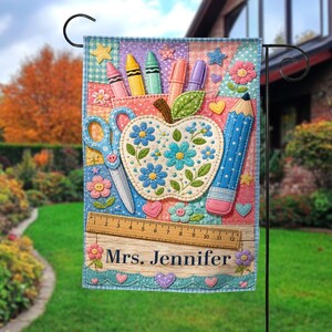 May include: A decorative garden flag featuring a colorful patchwork design with school-themed elements. The flag includes an apple, scissors, a pencil, crayons, and the name "Mrs. Jennifer". The flag is designed for outdoor use and is displayed on a black metal stand.