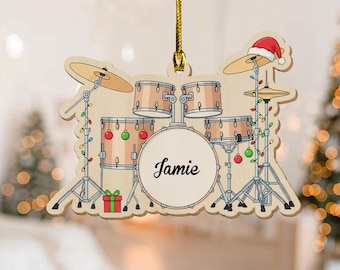 May include: Wooden Christmas ornament shaped like a drum set, with the name "Jamie" on the bass drum. The ornament features a Santa hat, Christmas lights, and a small gift box. The drum set is light brown with red and green accents.