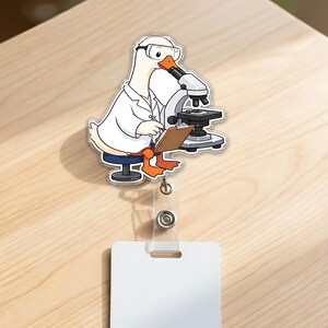 May include: A badge reel featuring a cartoon white duck wearing a lab coat and safety goggles, sitting on a stool while looking through a microscope and holding a clipboard. The duck has orange feet and an orange beak, with a clear plastic strap.