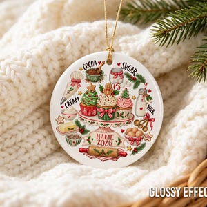 Pu&ograve; includere: Un ornamento rotondo in ceramica bianca con illustrazioni colorate di ingredienti da forno e cupcake su un'alzata. Il design include le scritte COCOA, SUGAR, CREAM, MILK e un nastro con il testo segnaposto NAME 2026.