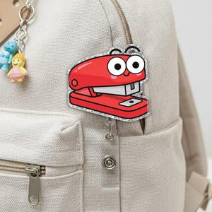 May include: A red stapler-shaped badge with a cartoon face and silver glitter trim is attached to a beige backpack. The badge has a smiling face with large white eyes and a silver staple. A small keychain with cartoon figures hangs from the backpack zipper.