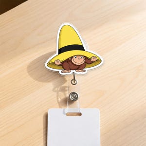 May include: A badge reel featuring a brown monkey character wearing a large yellow hat with a black band. The character is depicted with hands holding the brim of the hat. The badge reel includes a clear plastic strap and a white rectangular card holder.