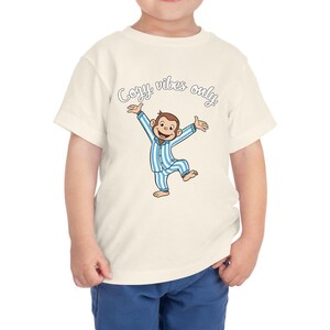 Op de afbeelding: Cr&egrave;me peuter t-shirt met een cartoon aap in blauw en wit gestreepte pyjama met uitgestrekte armen. De tekst "Cozy vibes only" staat in een scriptletter boven de aap.