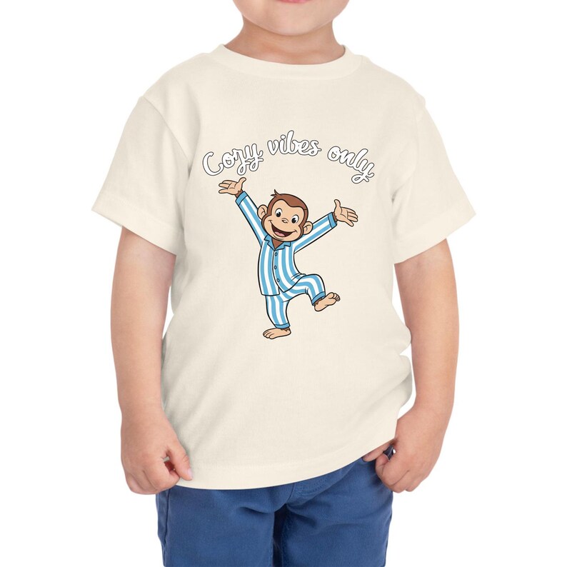 Op de afbeelding: Cr&egrave;me peuter t-shirt met een cartoon aap in blauw en wit gestreepte pyjama met uitgestrekte armen. De tekst "Cozy vibes only" staat in een scriptletter boven de aap.