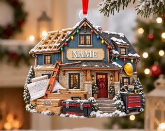 May include: A house-shaped Christmas ornament featuring a blue exterior, a red front door, and snow-covered roof. The structure displays construction elements including a ladder, blueprints, a toolbox, and the text NAME and 202X.