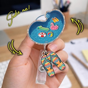 May include: A badge reel with a pool design, featuring a pink flamingo, a beach ball, and a green pool float. The reel also has a pair of colorful flip-flops attached. The text "Shake me!" is written in a script font.