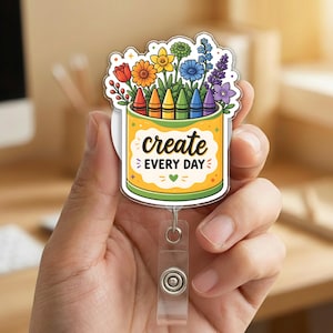 May include: A colorful badge reel featuring a crayon box design with the words "create EVERY DAY". The box is filled with crayons and a variety of flowers in rainbow colors. The reel has a clear plastic retractable cord and a metal clip.