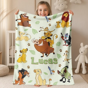 May include: A light green blanket featuring cartoon characters from The Lion King. The blanket has images of Simba, Timon, Pumbaa, and other characters. The name "Lucas" is printed in green. The blanket is decorated with paw prints and leaf designs.
