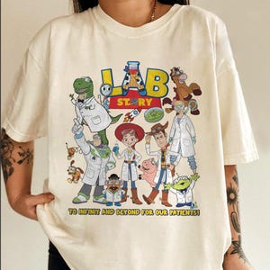 May include: Cream-colored t-shirt featuring a colorful graphic of cartoon characters in lab coats. The design includes the text "LAB STORY" and the phrase "TO INFINITY AND BEYOND FOR OUR PATIENTS!"