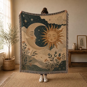 May include: A woven throw blanket featuring a celestial design with a large crescent moon and a radiant sun face. The scene includes stylized clouds, stars, and botanical elements in shades of cream, dark blue, and muted orange with fringed edges.