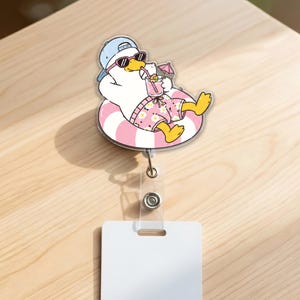 May include: A badge reel featuring a white duck wearing a light blue baseball cap and black sunglasses, lounging in a pink and white striped inflatable ring while holding a pink drink with a small umbrella, attached to a clear plastic strap.