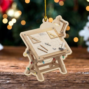 May include: Wooden Christmas ornament shaped like a drafting table, complete with a miniature lamp, pencils, and blueprints. The ornament is decorated with colorful Christmas lights and the name "Daniel" is visible on the blueprint. The ornament is hanging from a gold string.