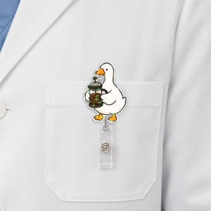 Puede incluir: Un portatarjetas de identificaci&oacute;n con forma de pato blanco, pico y patas naranjas, que sostiene una cafetera de &eacute;mbolo marr&oacute;n con destellos verdes. El personaje de acr&iacute;lico est&aacute; unido a un cord&oacute;n retr&aacute;ctil transparente y un clip met&aacute;lico.
