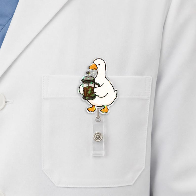 Puede incluir: Un portatarjetas de identificaci&oacute;n con forma de pato blanco, pico y patas naranjas, que sostiene una cafetera de &eacute;mbolo marr&oacute;n con destellos verdes. El personaje de acr&iacute;lico est&aacute; unido a un cord&oacute;n retr&aacute;ctil transparente y un clip met&aacute;lico.