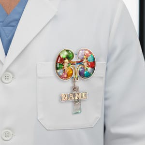 May include: A colorful kidney-shaped badge reel with a silver-tone metal clip. The badge features a mosaic design with various colors. The name tag below is in a gold color. The badge is attached to a white lab coat.