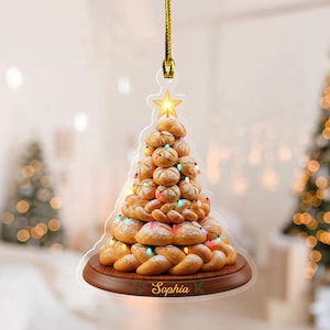 May include: A Christmas tree ornament shaped like a tree made of bread, decorated with string lights and a gold star. The ornament sits on a wooden base with the name "Sophia" printed on it. A gold string is attached for hanging.