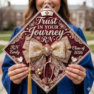 May include: A maroon graduation cap decorated with a white nurse's hat, a gold and lace bow, and a stethoscope. The cap reads "Trust in Your Journey RN" and includes the name "RN Emily" and "Class of 2026".