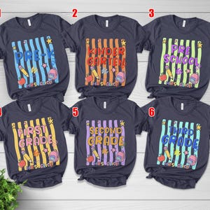 May include: Six navy blue t-shirts with school grade level designs. Each shirt features a vertical striped background with text indicating the grade level, along with school-themed graphics like pencils, apples, and backpacks.