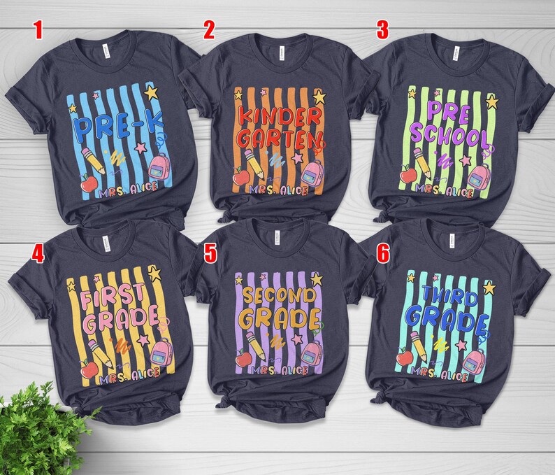 May include: Six navy blue t-shirts with school grade level designs. Each shirt features a vertical striped background with text indicating the grade level, along with school-themed graphics like pencils, apples, and backpacks.