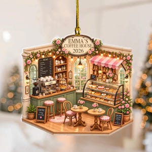 May include: A flat, hanging ornament shaped like a coffee shop interior with the text EMMA'S COFFEE HOUSE 2026. It features a green counter, a menu board, a pastry display case, two small round tables with chairs, and pink floral decorations.