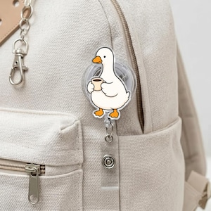 Puede incluir: Un portatarjetas de acr&iacute;lico blanco con una ilustraci&oacute;n de dibujos animados de un pato blanco con pico y patas naranjas, sosteniendo una peque&ntilde;a taza de caf&eacute; marr&oacute;n. El portatarjetas est&aacute; sujeto a una mochila de lona beige con un clip plateado.